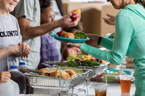 People serving food at a volunteer event, with trays of food and beverages.
