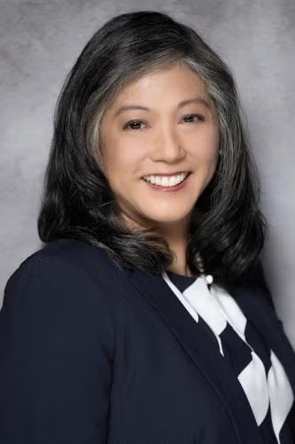 A smiling woman with gray hair in a dark jacket and white blouse against a gray backdrop.