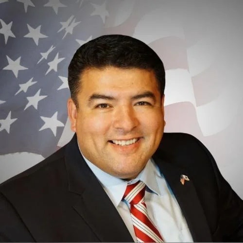 A smiling man in a suit with a striped tie before an American flag backdrop.