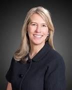 A person with long blonde hair, wearing a dark jacket, smiling, set against a dark background.