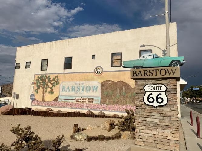 A mural of Barstow on Route 66, with a vintage car model on a signpost under a partly cloudy sky.