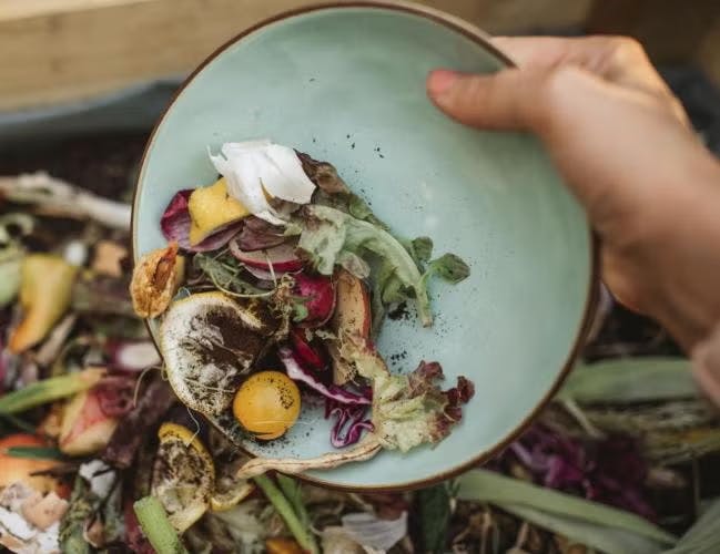 A hand holding a bowl with vegetable scraps and compostable waste, pouring them into a compost bin.