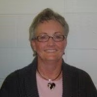 A person wearing glasses, a necklace, and a dark sweater, smiling in front of a light-colored wall.