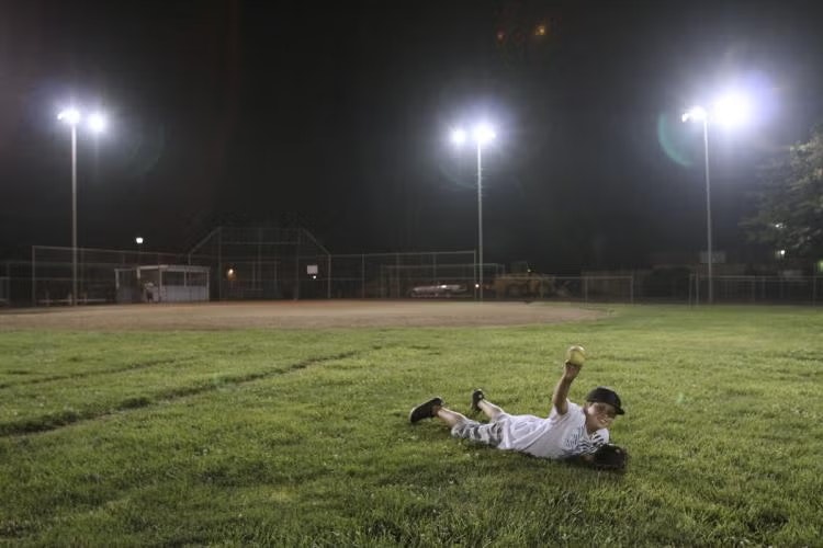 A child in a baseball uniform lies on grass under floodlights holding a baseball.