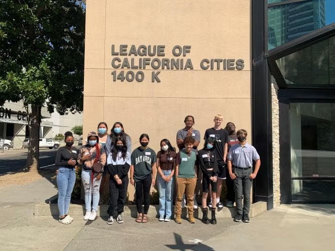 A group of people stand in front of a building with a sign reading "League of California Cities 1400 K."