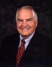 A smiling elderly man in a suit with a polka dot tie against a dark background.
