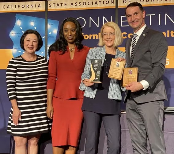 Four people smiling, two holding awards, in front of a California State Association of Counties backdrop.
