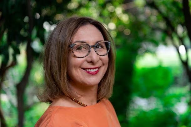 A smiling person with glasses and shoulder-length hair, wearing an orange top, with a green leafy background.