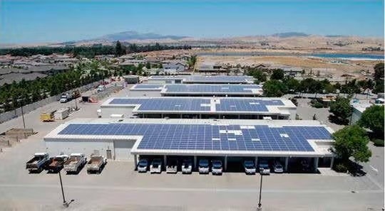Rooftops covered with solar panels on industrial buildings, trucks parked below, with a landscape of hills and greenery in the background.