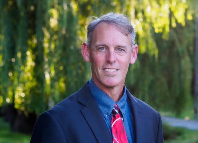 A person wearing a suit and red tie, standing outdoors with trees in the background.