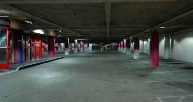 An empty parking garage with pink and white support columns, a flat ceiling, and some red entrance doors on one side.
