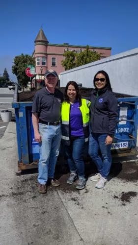 Three people posing in front of a blue dumpster on a sidewalk with a building and tree in the background.
