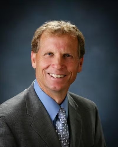 A smiling person in a suit, blue shirt, and patterned tie against a dark background.