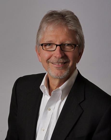 Portrait of a smiling man with grey hair and glasses, wearing a black blazer and white shirt against a grey backdrop.