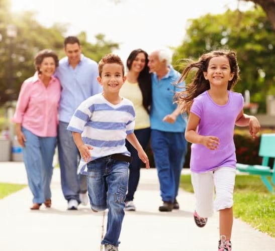 Two children running in a park, smiling adults walking behind them.
