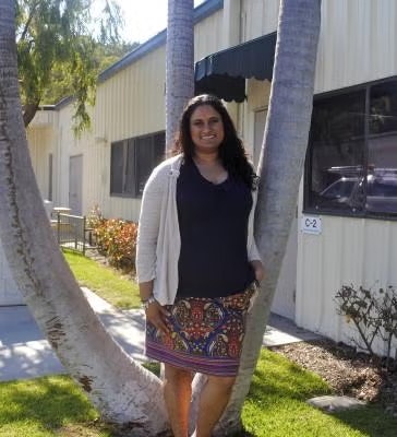 A person standing outside in front of a building, next to trees, wearing a navy top, patterned skirt, and light cardigan.