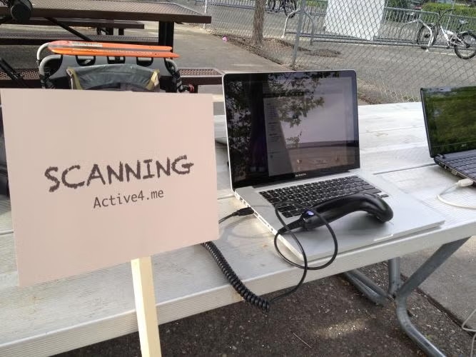A laptop with a barcode scanner on a picnic table beside a sign reading "SCANNING Active4.me".