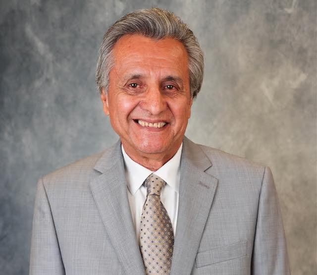 A man in a light gray suit and tie smiling against a gray background.