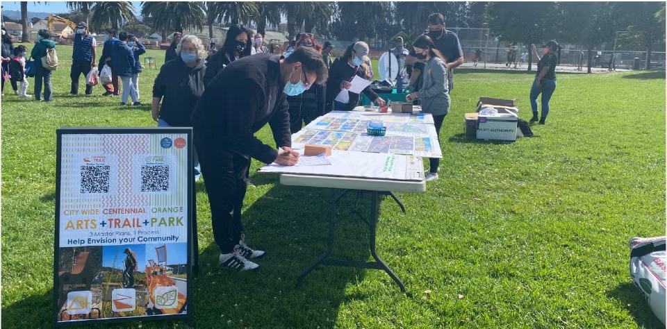 People participating in a community event outdoors, with a table, maps, and a sign about an arts and trail park project.