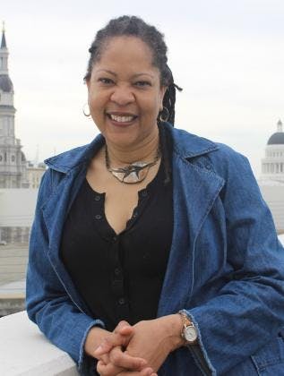 A smiling person with braided hair wearing a denim jacket and black top, standing outdoors with buildings in the background.