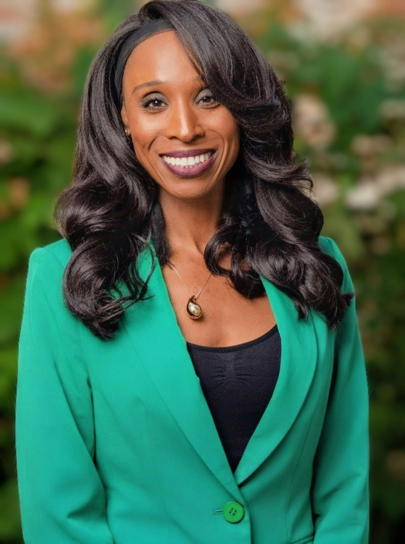A smiling individual with long, curly hair wears a green blazer over a black top, set against a blurred natural background.