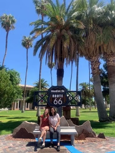 Two people sitting on a bench in front of a Route 66 sign surrounded by palm trees in a sunny park.