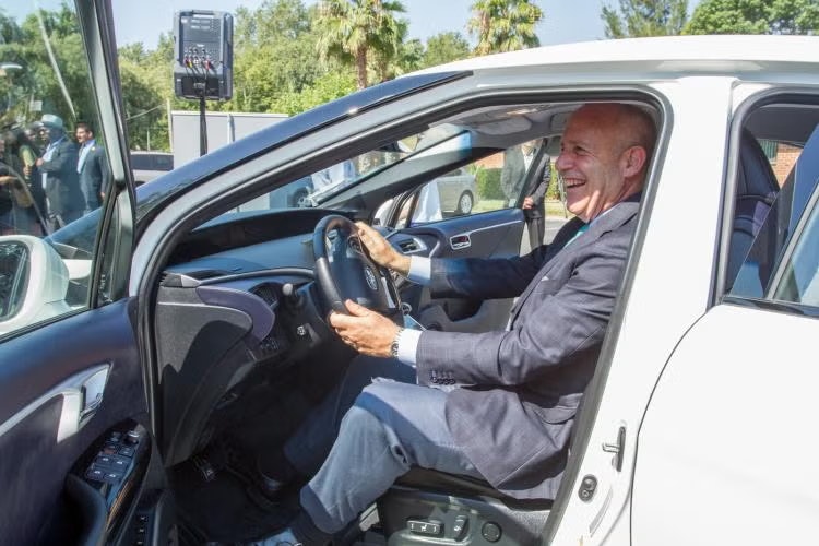 A person in a suit sits smiling in the driver's seat of a white car with the door open.