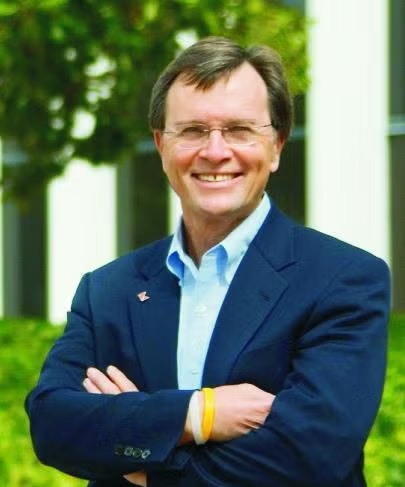 A person in a suit with arms crossed, smiling outdoors with greenery in the background.
