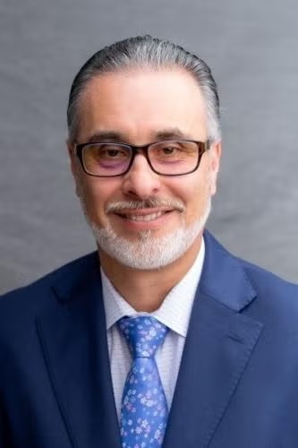 A man wearing glasses and a blue suit with a patterned tie, posing for a portrait.