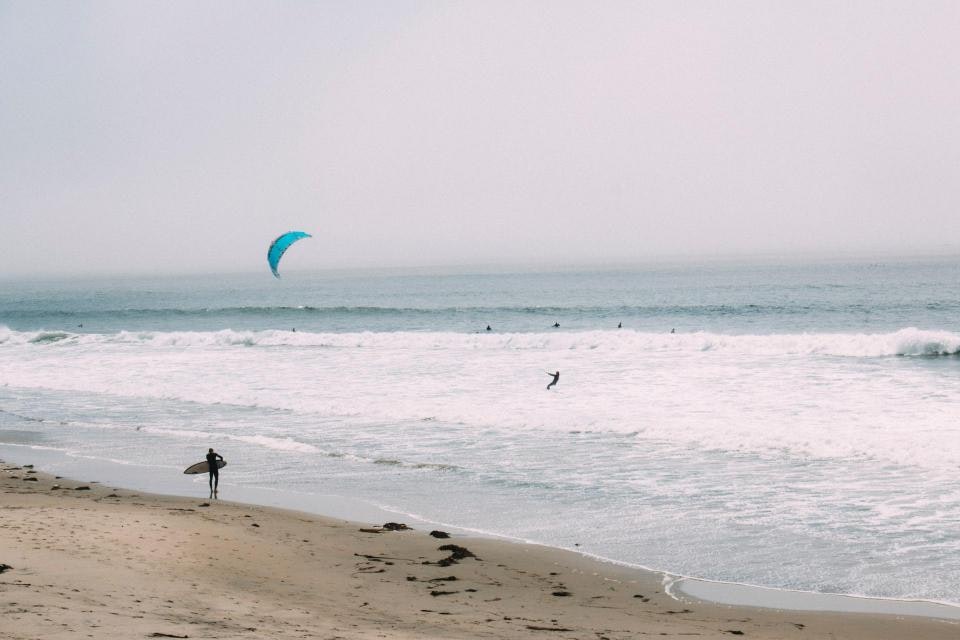 A beach scene with a surfer holding a board, a kite surfer in the water, and waves crashing onto the shore.