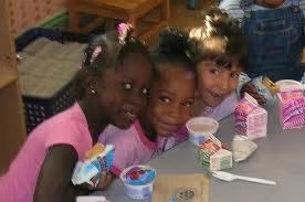 Three children smiling at a table with milk cartons, yogurt, and snacks.