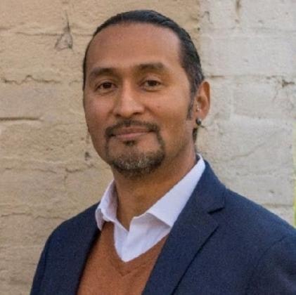 A person wearing a dark blazer, white shirt, and brown sweater in front of a textured wall.