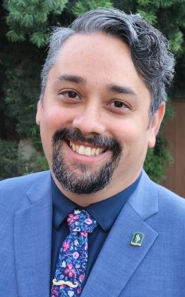 A smiling man with a beard, wearing a blue suit, patterned tie, and lapel pin, with greenery in the background.