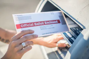 A hand is inserting an "Official Ballot Enclosed" envelope into a mailbox.