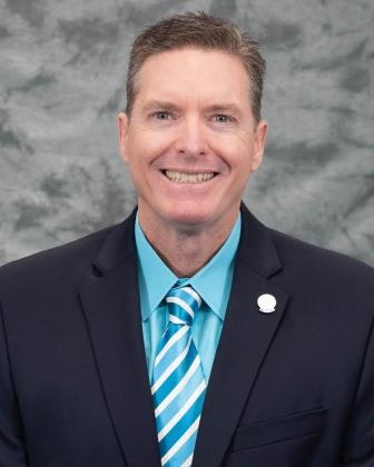 A person in a suit and tie, smiling against a gray backdrop.