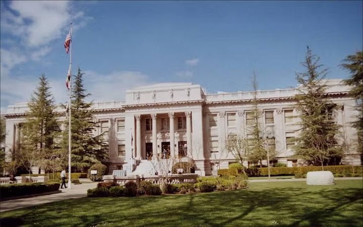 A large neoclassical building with columns, surrounded by trees and a garden, featuring a flagpole with the American flag.