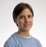 A person with short brown hair wearing a blue sweater, smiling against a plain background.