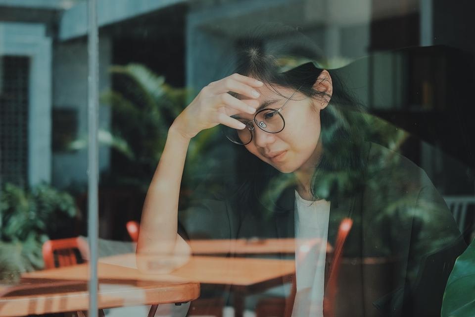 A person with glasses sits behind a window, resting their forehead on their hand, looking thoughtful or concerned.