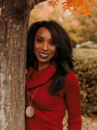 A person in a red sweater smiling beside a tree with autumn leaves.