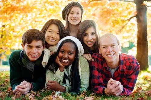 A group of six smiling people laying on grass with autumn trees in the background.