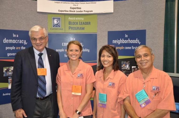Four people posing at a Cupertino Block Leader Program event with informational banners in the background.
