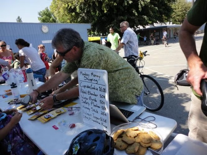 A man is selling items and snacks at an outdoor event. A sign lists prices for cookies, a pedal blender, and more. Bikes and people are around.