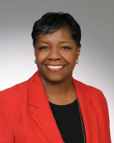 A person smiling, wearing a red blazer and a black top, with short hair and earrings, against a neutral background.