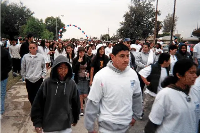 A large group of people walking down a street, some wearing hoodies and t-shirts, with a balloon arch in the background.