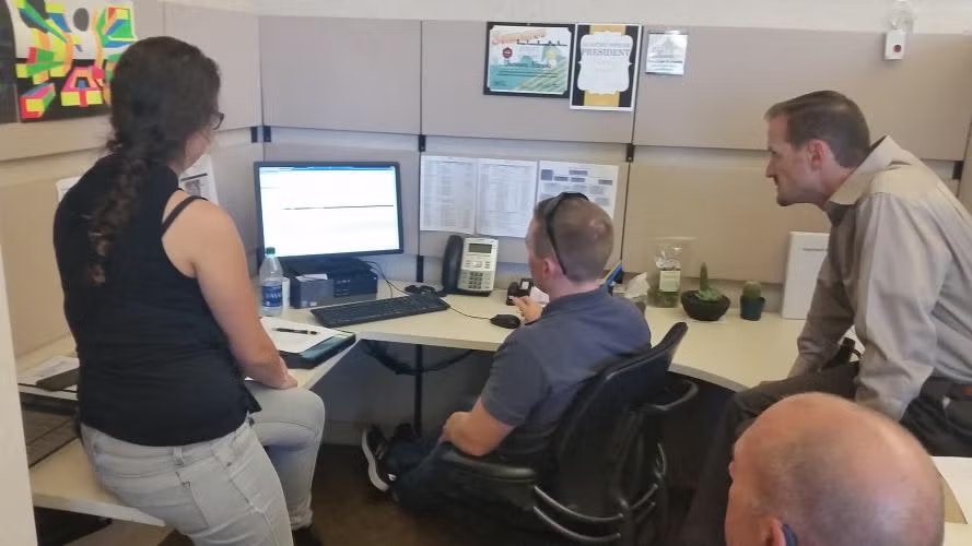 Four people in an office cubicle gathered around a computer screen, engaged in discussion.