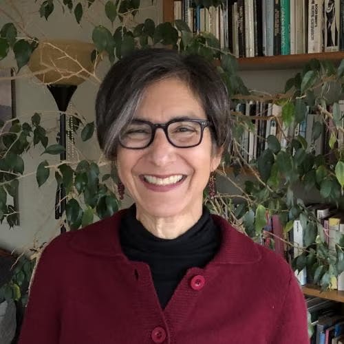 A person with glasses smiling in front of a bookshelf and plants, wearing a maroon top.