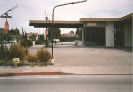 A deserted street corner with a modernist building, street signs, and minimal landscaping under a cloudy sky.