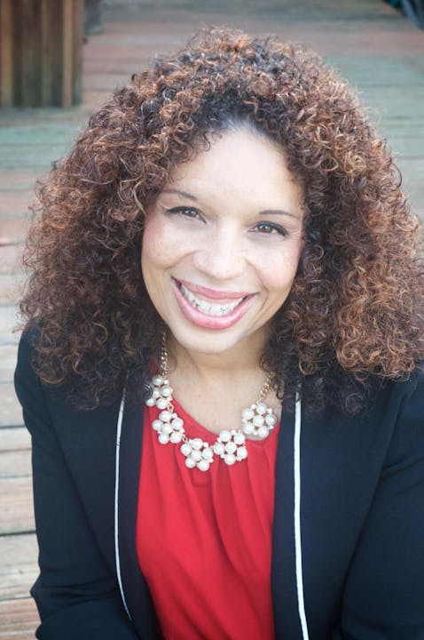 Person smiling, with curly hair, wearing a necklace and a red top with a black blazer.