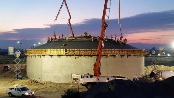 Construction workers on a large cylindrical structure using cranes at sunset.