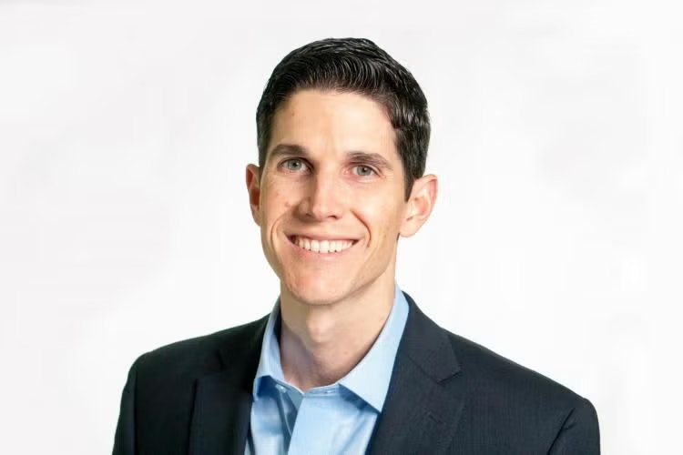 A person smiling, wearing a dark suit and blue collared shirt, against a plain light background.